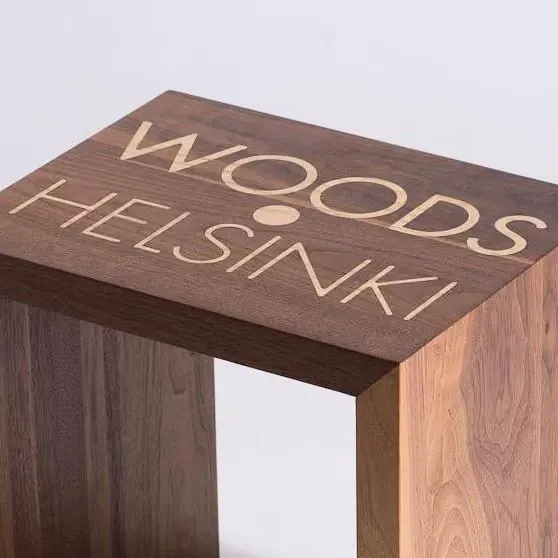 Woods Helsinki -tunnus vaaleasta puusta upotettuna pähkinäpuiseen jakkaraan